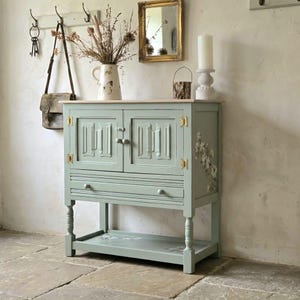 May include: A sage green wooden cabinet featuring two doors with gold-toned hinges, a single drawer with two knobs, and a lower shelf. The cabinet is decorated with floral motifs on the side and bottom shelf, standing on four turned legs.