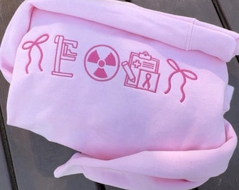 May include: A light pink hoodie with embroidered pink graphics. The design includes a bow, the letters "KE", a radiation symbol, a clipboard with a plus sign, and a ribbon. The hoodie is folded on a wooden surface.