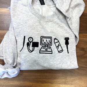 Peut inclure: Un sweat-shirt gris clair avec des graphiques noirs d'équipements médicaux, dont un stéthoscope, un tensiomètre et un appareil à ultrasons. Le sweat-shirt est plié sur une surface en bois.