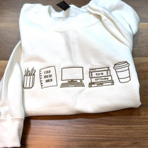 May include: Cream-coloured sweatshirt featuring embroidered brown line art of office supplies, a computer, books, and a coffee cup. The sweatshirt also features numbers and the text "ICD-10" and "CPT Coding".