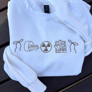 Könnte beinhalten: Weißes Sweatshirt mit Rundhalsausschnitt und braunen, gestickten medizinischen Symbolen, darunter ein CT-Scanner, ein Strahlungssymbol und eine medizinische Tabelle. Das Sweatshirt hat lange Ärmel und einen gerippten Kragen und Bündchen. Das Design ist auf der Brust zentriert.