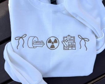 Puede incluir: Sudadera blanca de cuello redondo con símbolos médicos bordados en marrón, incluyendo un escáner CT, un símbolo de radiación y una tabla médica. La sudadera tiene mangas largas y cuello y puños acanalados. El diseño está centrado en el pecho.