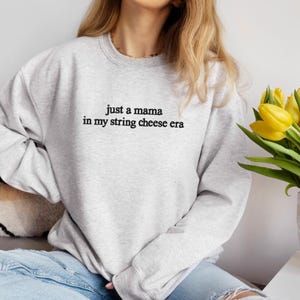 May include: A light gray sweatshirt with the text "just a mama in my string cheese era" in black. The sweatshirt is a crew neck style. Yellow tulips are visible in the background.