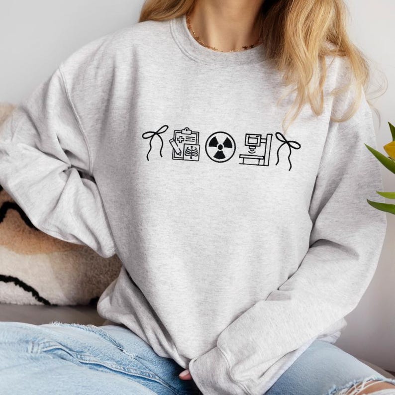 May include: Light gray sweatshirt with black line art of medical symbols, including a clipboard with an X-ray, a radiation symbol, and a medical machine. The sweatshirt has a crew neck and long sleeves.