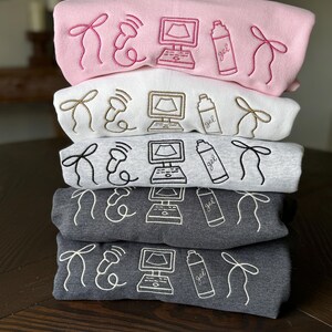 May include: A stack of five sweatshirts in various colors: pink, white, gray, and dark gray. Each sweatshirt features embroidered medical-themed designs, including a bow, an ultrasound probe, a computer screen, and a gel bottle. The designs are in a contrasting color to the sweatshirt.