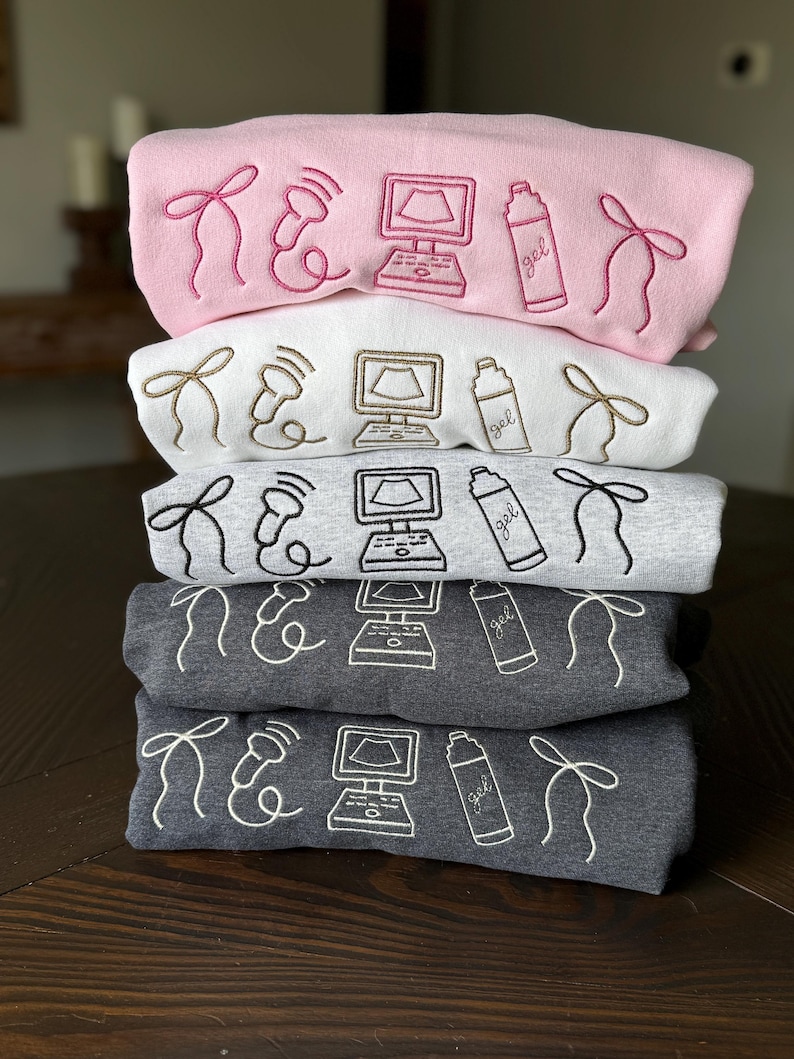 May include: A stack of five sweatshirts in various colors: pink, white, gray, and dark gray. Each sweatshirt features embroidered medical-themed designs, including a bow, an ultrasound probe, a computer screen, and a gel bottle. The designs are in a contrasting color to the sweatshirt.