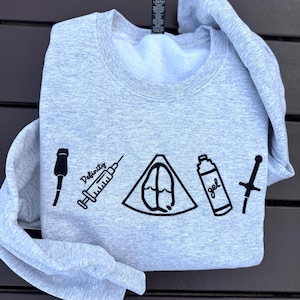Puede incluir: Una sudadera gris claro con diseños gráficos negros. Los diseños incluyen un micrófono, una jeringa con la palabra "Definitely", un triángulo con un diseño en el interior, una botella de gel y una espada.
