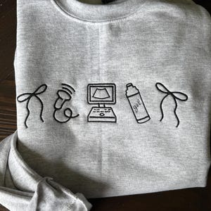 May include: A heather gray sweatshirt with black embroidered medical-themed graphics. The design includes a stethoscope, a computer monitor, a bottle of gel, and decorative elements. The sweatshirt is made of a soft material.
