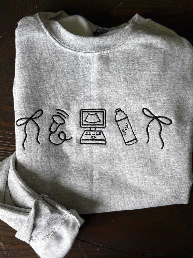 May include: A heather gray sweatshirt with black embroidered medical-themed graphics. The design includes a stethoscope, a computer monitor, a bottle of gel, and decorative elements. The sweatshirt is made of a soft material.