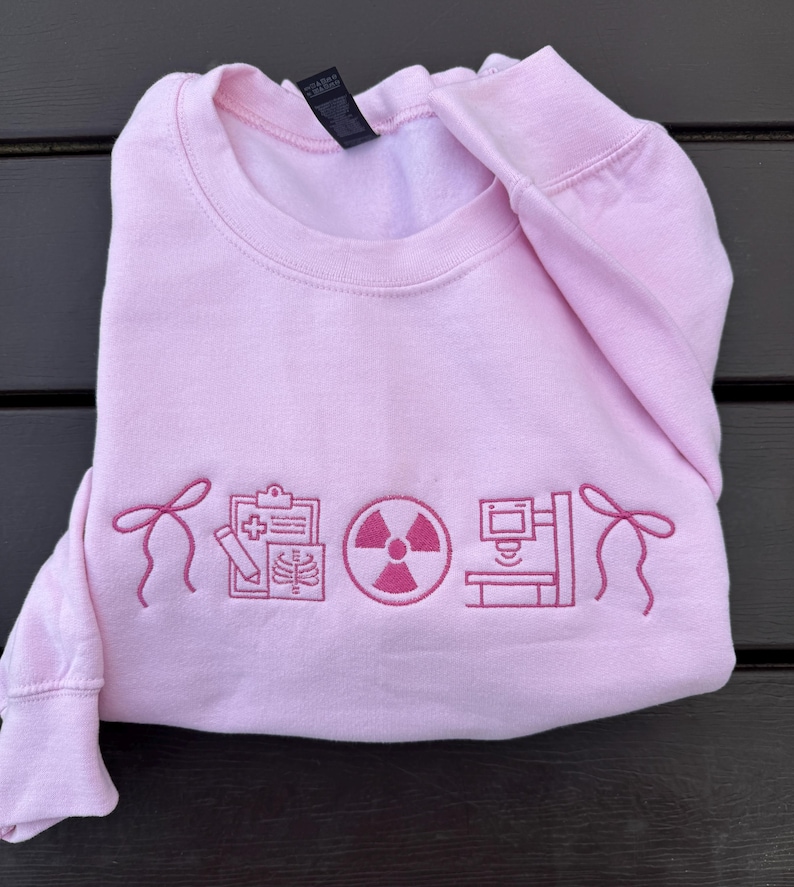 May include: A light pink sweatshirt with embroidered medical-themed graphics. The design includes a bow, a clipboard with an X-ray, a radiation symbol, and a medical imaging machine. The sweatshirt is folded neatly.