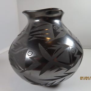 May include: A black ceramic vase with a glossy finish. The vase features a repeating pattern of stylized fish and geometric shapes etched into the surface. The date 01/01/2000 is visible on the bottom right.