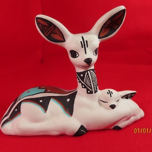 May include: A white ceramic figurine of a mother deer lying down with a small fawn resting against its side. The deer features black, brown, and turquoise geometric patterns on its body and ears. The date 01/01/2000 is printed in the bottom corner.