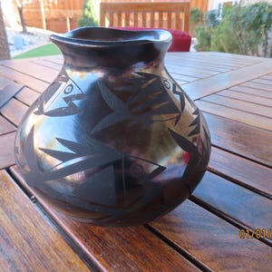 May include: A black pottery vase with a glossy finish. The vase features a design of stylized fish and geometric shapes. The date "01/01/2000" is visible on the bottom right. The vase is sitting on a wooden table.