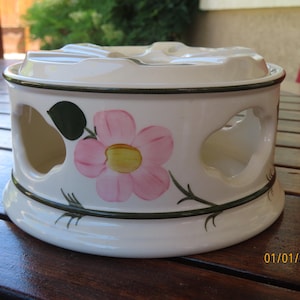 May include: Cream-colored ceramic ashtray with a floral design. The ashtray features a pink flower with a yellow center and green leaves. It has a round shape with cut-out sides and a dark green trim.