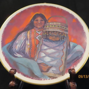 May include: A ceramic plate with a colorful painting of a Native American woman holding a baby. The woman is wearing a red and blue patterned dress and the baby is wearing a white and blue patterned blanket. The background is a red and orange sunset.
