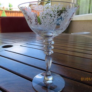 May include: A clear, etched glass champagne coupe with a wide bowl and a decorative floral pattern. The stem features a series of stacked rings and a fluted design. The coupe sits on a round base, reflecting light. The glass is on a wooden table.