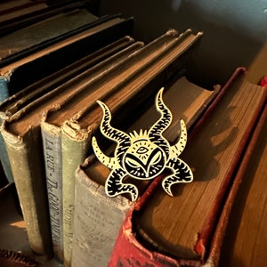May include: A gold-colored pin with a black and white design of a creature with horns, an eye, and a mouth. The pin is sitting on top of a stack of old books.