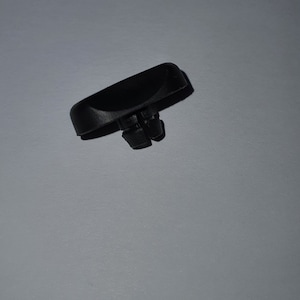 Könnte beinhalten: Ein kleines, schwarzes Kunststoffteil mit einer rechteckigen Oberseite und zwei kleinen Vorsprüngen an der Unterseite. Die Komponente scheint Teil eines größeren Mechanismus oder Geräts zu sein, möglicherweise ein Knopf oder Schalter.