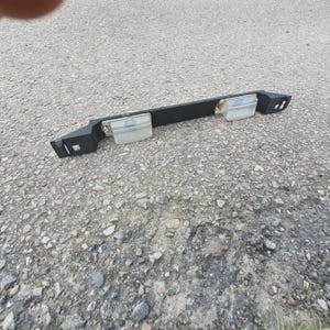 May include: A black car number plate light assembly with two clear, rectangular light housings. The assembly is rectangular with angled ends and rests on a grey surface.