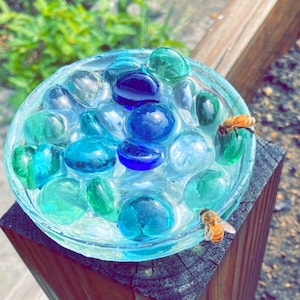 May include: A clear glass bowl filled with blue and green glass marbles. Two bees are perched on the rim of the bowl.