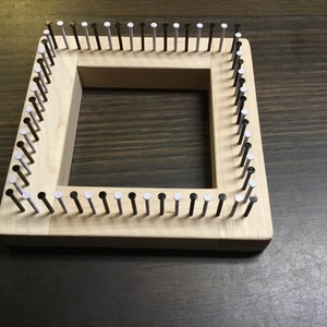 May include: A square wooden knitting loom with evenly spaced nails around the perimeter. The nails alternate between black and white heads. The loom is made of light-colored wood and is designed for yarn crafts.