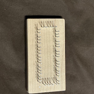 May include: A rectangular wooden board with a light brown finish. Small silver nails are hammered into the wood in a rectangular pattern, creating a frame-like design. The nails are evenly spaced.