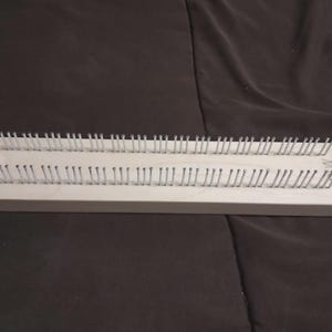 May include: A rectangular wooden knitting loom with evenly spaced metal pegs. The loom is light-colored and has two rows of pegs, ideal for creating knitted items. The pegs are silver and stand upright.