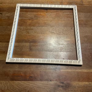 May include: A square, wooden weaving loom with small metal pins along the edges. The loom is made of light-colored wood and is set against a dark wood surface. The pins are evenly spaced and create a grid-like appearance, ideal for textile crafts.