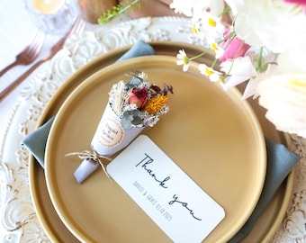 May include: A small bouquet of dried flowers wrapped in a paper cone with a tag that says "Coupled & Grace". The bouquet is sitting on a gold plate with a white thank you card that says "Thank you Daniel & Grace 03.18.2023".