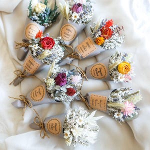 以下が含まれることがあります： グレーの紙に包まれた、ドライフラワーの小さなブーケの詰め合わせ。茶色の紙のタグには、「ダニエル＆グレイス」など、カップルの名前が書かれています。