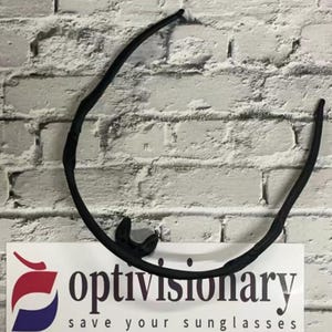 May include: Black plastic sunglasses frame with a flexible, curved design. The frame is shown against a white brick wall.