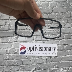 Puede incluir: Gafas de sol sin montura negras de forma rectangular. Las gafas se sostienen en una mano contra una pared de ladrillo blanco. El texto "optivisionary save your sunglasses" es visible en la esquina inferior derecha de la imagen.