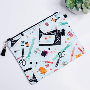May include: A blue zippered pouch with a black zipper and a black tassel. The pouch has a white background with a colorful pattern of sewing tools, including a sewing machine, spools of thread, scissors, and needles.