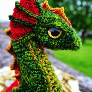 K&ouml;nnte beinhalten: Gr&uuml;ner geh&auml;kelter Drache mit roten und goldenen Akzenten. Der Drache hat ein gro&szlig;es, goldenes Auge und einen stacheligen, roten Kamm. Der Drache sitzt auf einem Haufen Goldm&uuml;nzen.