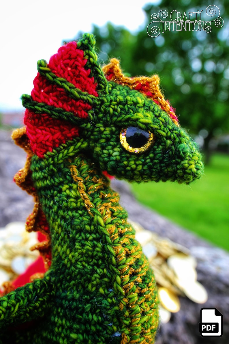 K&ouml;nnte beinhalten: Gr&uuml;ner geh&auml;kelter Drache mit roten und goldenen Akzenten. Der Drache hat ein gro&szlig;es, goldenes Auge und einen stacheligen, roten Kamm. Der Drache sitzt auf einem Haufen Goldm&uuml;nzen.