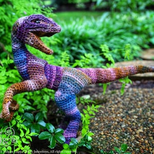 May include: A crocheted dinosaur toy in shades of purple, blue, and brown, standing on a bed of green leaves and gravel.
