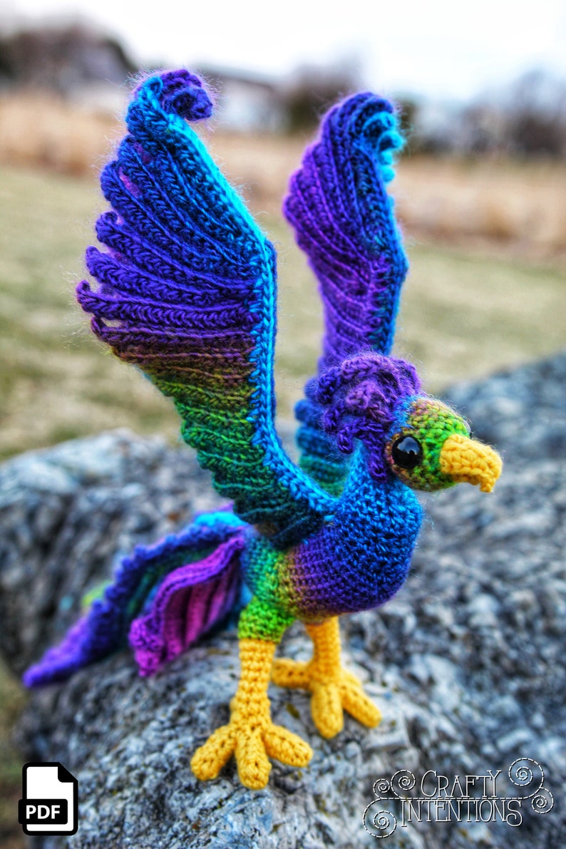 May include: A crocheted rainbow-colored phoenix with yellow legs and black eyes. The phoenix is standing on a gray rock.