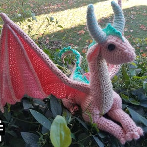 May include: A pink crocheted dragon with teal accents and wings. The dragon is sitting on a bed of green leaves.