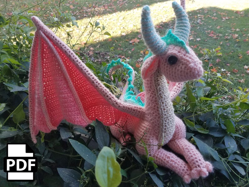 May include: A pink crocheted dragon with teal accents and wings. The dragon is sitting on a bed of green leaves.