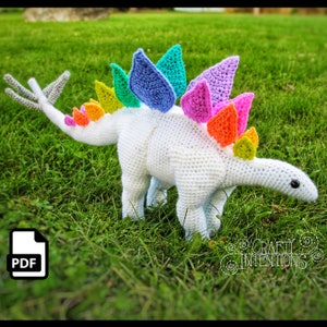 May include: A white crocheted dinosaur with a rainbow coloured spiky back. The dinosaur is standing on green grass. The image includes a PDF icon in the bottom left corner.