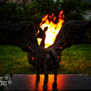 May include: Black crochet amigurumi sculpture of a mythical creature with wings, standing in front of a fire pit. The sculpture is in front of a black surface with the text "CRAFTY INTENTIONS" and a PDF icon.