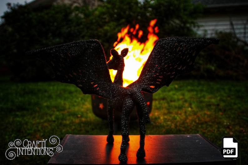 May include: Black crochet amigurumi sculpture of a mythical creature with wings, standing in front of a fire pit. The sculpture is in front of a black surface with the text "CRAFTY INTENTIONS" and a PDF icon.