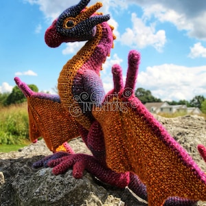May include: A crocheted dragon with orange, pink, and purple yarn. The dragon is sitting on a rock with a blue sky and white clouds in the background. The image includes the text "Crafty Intentions" and a PDF icon.
