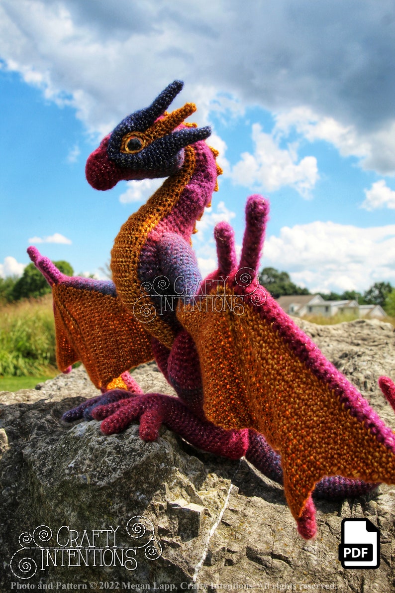 May include: A crocheted dragon with orange, pink, and purple yarn. The dragon is sitting on a rock with a blue sky and white clouds in the background. The image includes the text "Crafty Intentions" and a PDF icon.