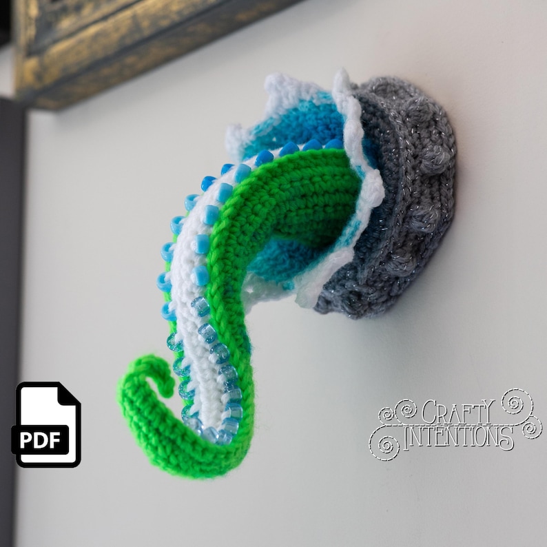 May include: A crocheted octopus tentacle in shades of green, white, and blue, with a gray head. The tentacle is adorned with blue beads. The design is mounted on a white surface. The text "Crafty Intentions" is visible.