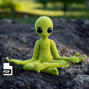 May include: A green crocheted alien doll sitting in a meditative pose with its arms outstretched. The doll has large black eyes and a simple, rounded body.
