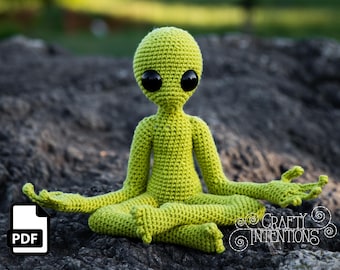 May include: A green crocheted alien doll sitting in a meditative pose with its arms outstretched. The doll has large black eyes and a simple, rounded body.
