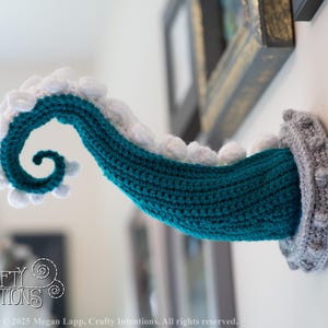 May include: A teal and white crocheted tentacle emerges from a gray, textured wall mount. The tentacle is curled and detailed, with white, scalloped edges. The design is a whimsical, handmade craft project. The image includes the text "Crafty Intentions" and a PDF icon.