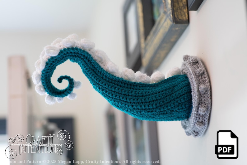 May include: A teal and white crocheted tentacle emerges from a gray, textured wall mount. The tentacle is curled and detailed, with white, scalloped edges. The design is a whimsical, handmade craft project. The image includes the text "Crafty Intentions" and a PDF icon.
