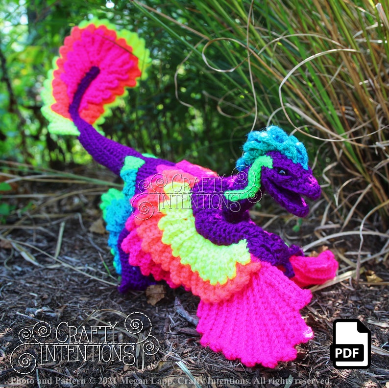 May include: A crocheted rainbow dragon with a purple body, green and blue head, and a pink tail. The dragon is standing on a bed of brown mulch and green grass.